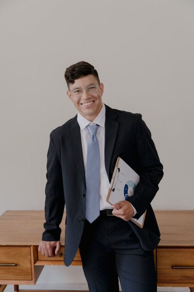 Confident businessman in a black suit smiling, holding clipboard indoors.